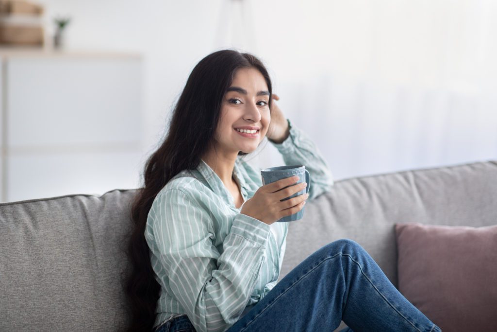 Portrait of young Indian woman having coffee break, sitting on sofa with cup of hot beverage at home. Lovely millennial lady relaxing on couch, enjoying lazy morning in living room