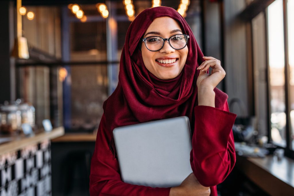 Pretty young islamic female holding a laptop while at a cafe. Smiling businesswoman in hijab at coffee shop.