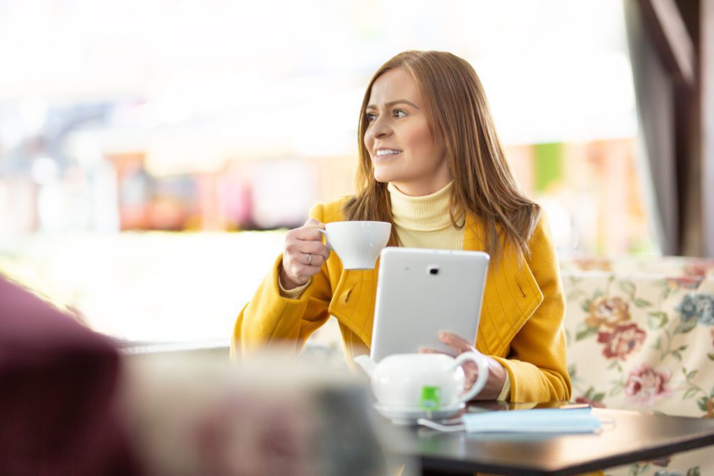 Woman wearing turtleneck drinks a cup of tea holding a tablet in a cafe.